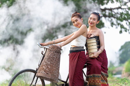 Beautiful girls wearing Thai traditional dress in the countryside takes her bicycle back from the rice fields and random catch fish on bike. One holding a vintage tiffin container.Portrait smileの写真素材