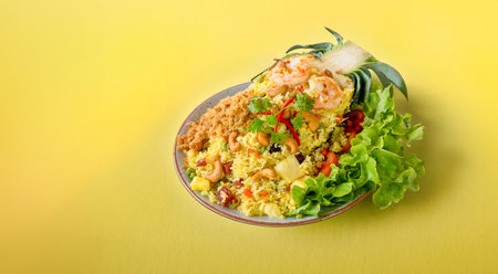 Freshly prepared pineapple fried rice served inside of half a pineapple like boat on a plate on a yellow background. Thai pineapple stuffed with rice, shrimp, chicken and vegetables on the tableの写真素材