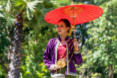 Asian beautiful lady wear a luxury national dress holding a red umbrella and Thai wooden old style house background. Young woman in Thai dress and Thai houseの写真素材