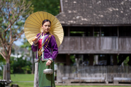 Asian beautiful lady wear a luxury national dress holding a yellow umbrella and Thai wooden old style house background. Young woman in Thai dress and Thai houseの写真素材