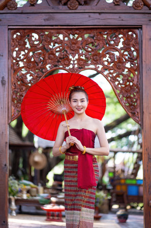 Beautiful Asian women wearing Thai traditional Lanna style dresses posing, holding a red traditional umbrella in ancient Thai house. Thai national costume. Travel conceptの写真素材