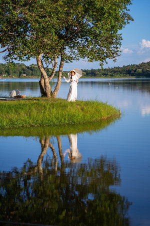 Beautiful woman in white edwardian style dress and white big hat standing in side lake and reflection. Woman in vintage blouse costume. Historical dresses, victorian or edwardian era style dressの写真素材