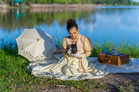 Charming and beautiful young Asian women in victorian or edwardian era holding retro camera and having an afternoon tea party in nature on the river bank, enjoy picnicking, reading book and sipping tea.の写真素材