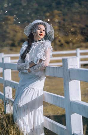 Beautiful woman in white edwardian style dress and white big hat standing in White fence in the pasture.Portrait of aristocratic girl.vintage blouse costume. Historical dresses, victorian or edwardianの写真素材