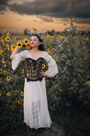 A beautiful woman in a beige dress stands in a field with blooming yellow sunflowers at sunset. Femlae model posing alone in middle of sunflower field. Morning or evening. Summertimeの写真素材