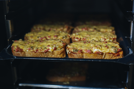 Hot toasted sandwiches with ham, cucumbers and mayonnaise cooked in a roaster and laid in rows on a black trayの写真素材