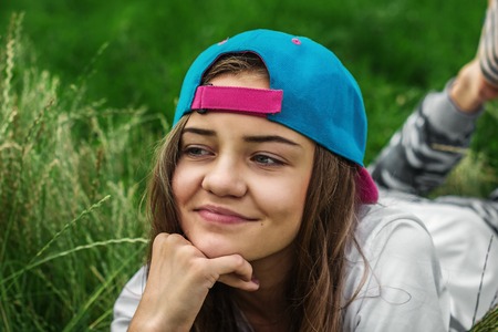 Beautiful white Caucasian girl in a baseball cap lies on a green grass on a summer day and looks...