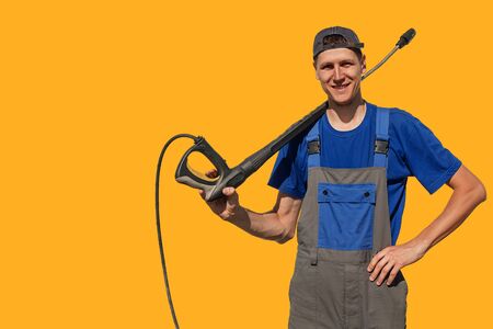 Tall slim guy with a gun for a car wash isolated on yellow background. A worker in coveralls looks at the camera and smiles. Background or layout with space for copyの写真素材