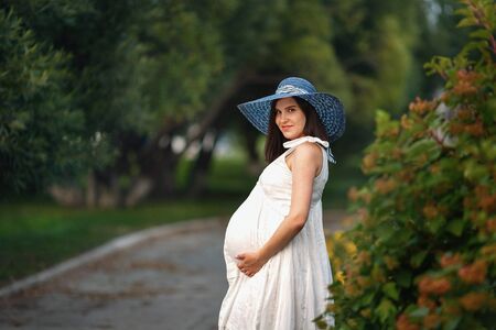Young beautiful pregnant girl of Caucasian appearance in white dress and hat in nature in the Park