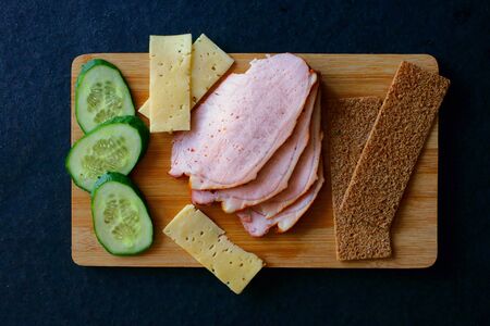 Slicing meat ham, cucumbers, cheese and ruddy breads on a wooden board. A look from above. Fresh and juicy breakfast or snackの写真素材