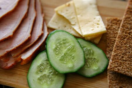 Slicing ham, cucumbers and cheese with ruddy thin breads lie on a wooden board.の写真素材