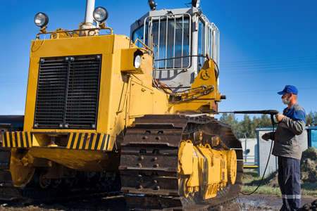 Car wash of heavy machinery and tractors. A man washes a yellow crawler tractor on the street on a sunny summer day.の写真素材
