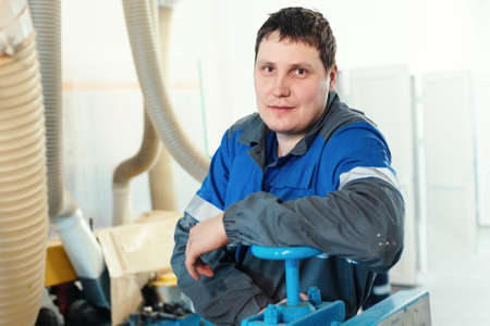 Portrait of a young worker in overalls at the workplace at the machine. A calm and relaxed white man of Caucasian appearance looks directly into the camera. Industrial background. Work in production.の写真素材