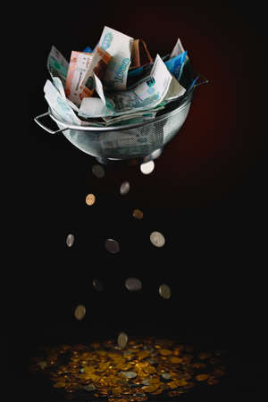 From a sieve with a pile of Russian paper rubles, coins fall into a large pile. The concept of inflation and economic poverty. Dark background.の写真素材