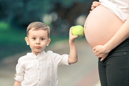 Portrait of a little boy next to a pregnant mother on a summer day in the park. The child holds an apple near the mother's big belly. Care and love in the family.の写真素材