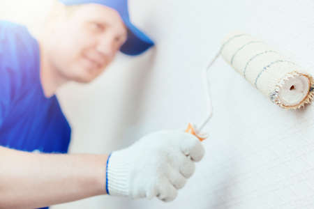 A painter in a cap and gloves paints the wall with a paint roller. Portrait of a young worker in...