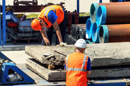 Two slingers unload concrete slabs on the street on a summer day. Workers in construction helmets and vests sling cargo. Work in an open warehouse.の写真素材