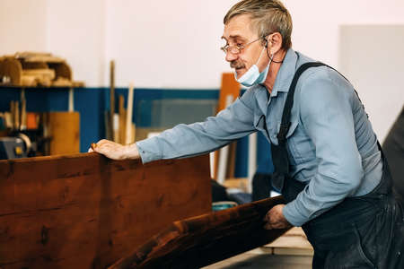 A gray-haired carpenter with glasses works with wood on a workbench in a carpentry shop. A pensioner...