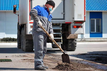 A male laborer in a medical mask and overalls works with a shovel and cleans the territory of the...