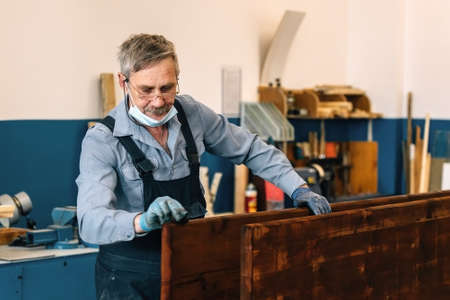 An elderly man paints wooden boards with dark varnish in a carpentry workshop. A pensioner in a medical mask earns a part-time job as a cabinetmaker. Physical laborの写真素材