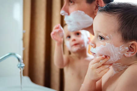 A man and two little boys with shaving foam on their faces look thoughtfully in the bathroom mirror. Father and sons fool around. Growing up and imitating adults.の写真素材