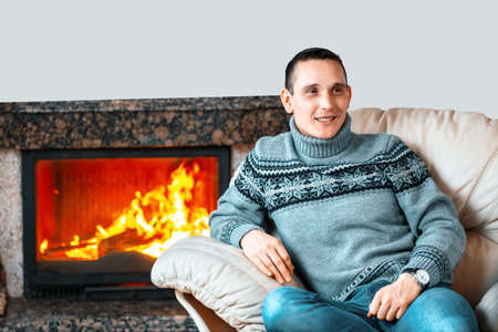 A young man in a knitted sweater sits in a leather chair against the background of a fireplace with fire and smiles. Home comfort and warmth from a luxurious fireplace. copy spaceの写真素材