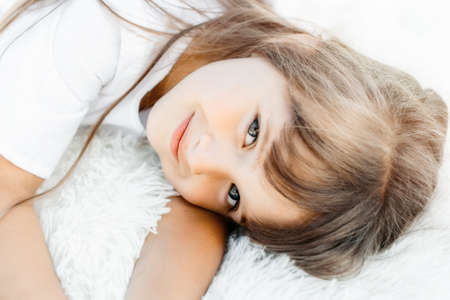 A portrait of a beautiful Caucasian girl lies on a white soft carpet with pile. Smile and joy on the childs face. Light tonality.の写真素材