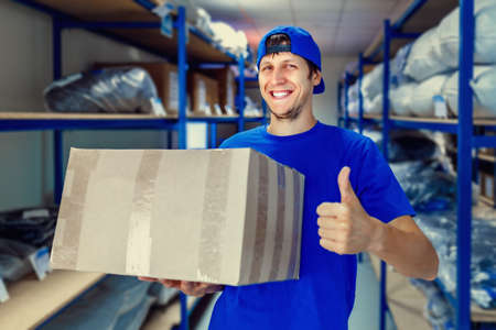 A young positive warehouse worker holds a cardboard box in his hands and smiles. The concept of storage and delivery of goods.の写真素材