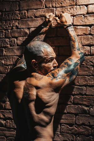 A criminal in tattoos stands against a brick wall with handcuffs on his hands. A dramatic portrait of a prisoner with a naked muscular body. Arrest for a crime.の写真素材