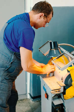 A Caucasian worker in uniform sharpens a part on a machine in a workshop. Manual processing of...
