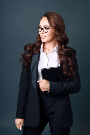 A young beautiful brunette woman in glasses and a business suit smiles and looks away. Studio portrait of a successful business lady on a gray background. Business Coachの写真素材