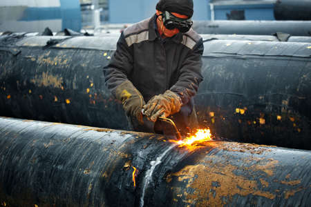 The welder cuts large metal pipes with ocetylene welding. A worker on the street cuts large-diameter...
