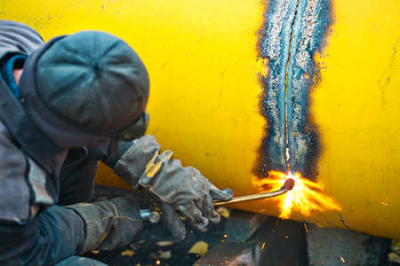 The welder cuts a large yellow pipe with acetylene welding for gasification. Disposal of old used metal pipes. Authentic workflow scene. Industrial background.の写真素材
