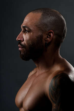 Brutal portrait of Latin American with short haircut and pumped muscles. Strong man with naked body looks away. Studio portrait on dark background close-up. Sweaty athletic body.の写真素材