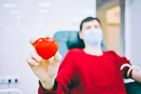 Man sits in chair and donates blood in medical clinic. Topic of donation. Donor holds plastic heart...