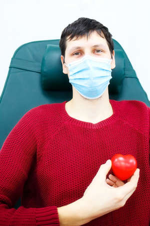 Young man in medical mask sits in chair and holds small heart in his hand. Grateful patient. Topic of health care and donation. Rescue of sick peopleの写真素材