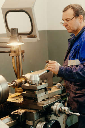 Turner in overalls works behind lathe in production hall. Processing of metal products. Industrial...