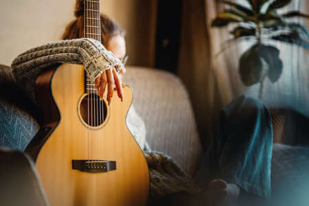 Young girl sits in room on couch with guitar. Teenager is hipster with musical instrument. Authentic scene.の写真素材