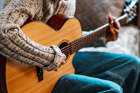 Young girl with long nails plays acoustic guitar at home. Teenager sits on couch in room and learns...