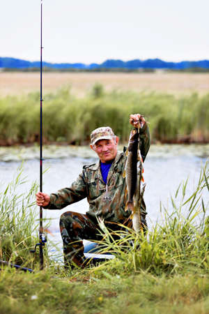 a fisherman with fishing rod shows catch of fish. Elderly man in camouflage sits by river or pond in reeds and shows caught pike. Outdoor activities during day.の写真素材