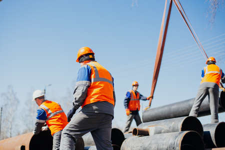 Group of slingers in construction helmets and vests on street unload metal pipes. Real scene....