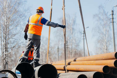 Slinger in helmet unloads metal pipes on construction site on sunny summer day. Real scene....