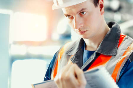 Young inspector or engineer in helmet and construction vest makes journal entries at production...