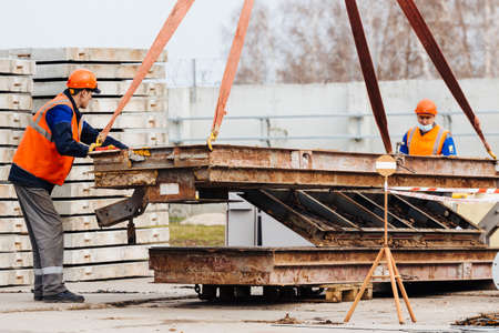 Slinger in helmet and vest controls unloading of metal structures on construction site. White handyman unloads load.の写真素材