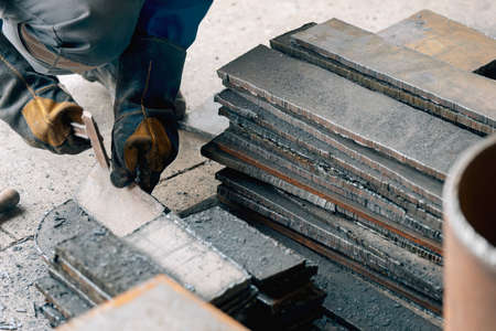 Worker in heavy industry or metallurgy beats scale off iron plates with hammer. Work with metal. Procurement of recyclables for production. Authentic workflow.の写真素材