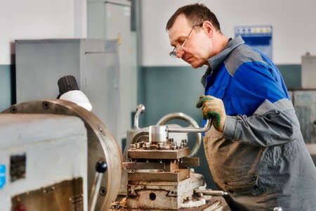 Elderly turner of 50-55 years old in overalls works at lathe in production shop. Processing of metal products. Real portrait of Caucasian worker. Production background.の写真素材