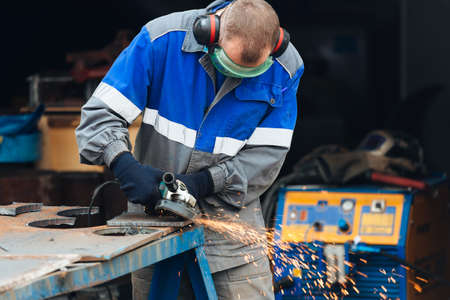 Real worker in working clothes grinds metal surface and sparks fly. Real portrait of person at work