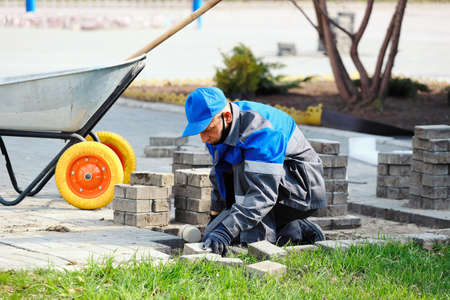 Bricklayer in work clothes sits on sidewalk and lays out paving slabs. Sight of working man in open air. Professional builder makes arrangement of territory summer day. Real scene.の写真素材