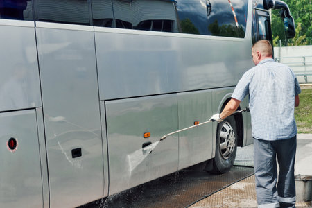 Car wash worker manually washes large bus with stream of water in open parking lot on summer day. Service for washing large equipment. Authentic scene workflow.の写真素材