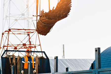 Slinger in helmet and vest unloads metal pipes and fittings from truck body on clear day.. Production background. Authentic workflow on construction site. Slinger at work..の写真素材